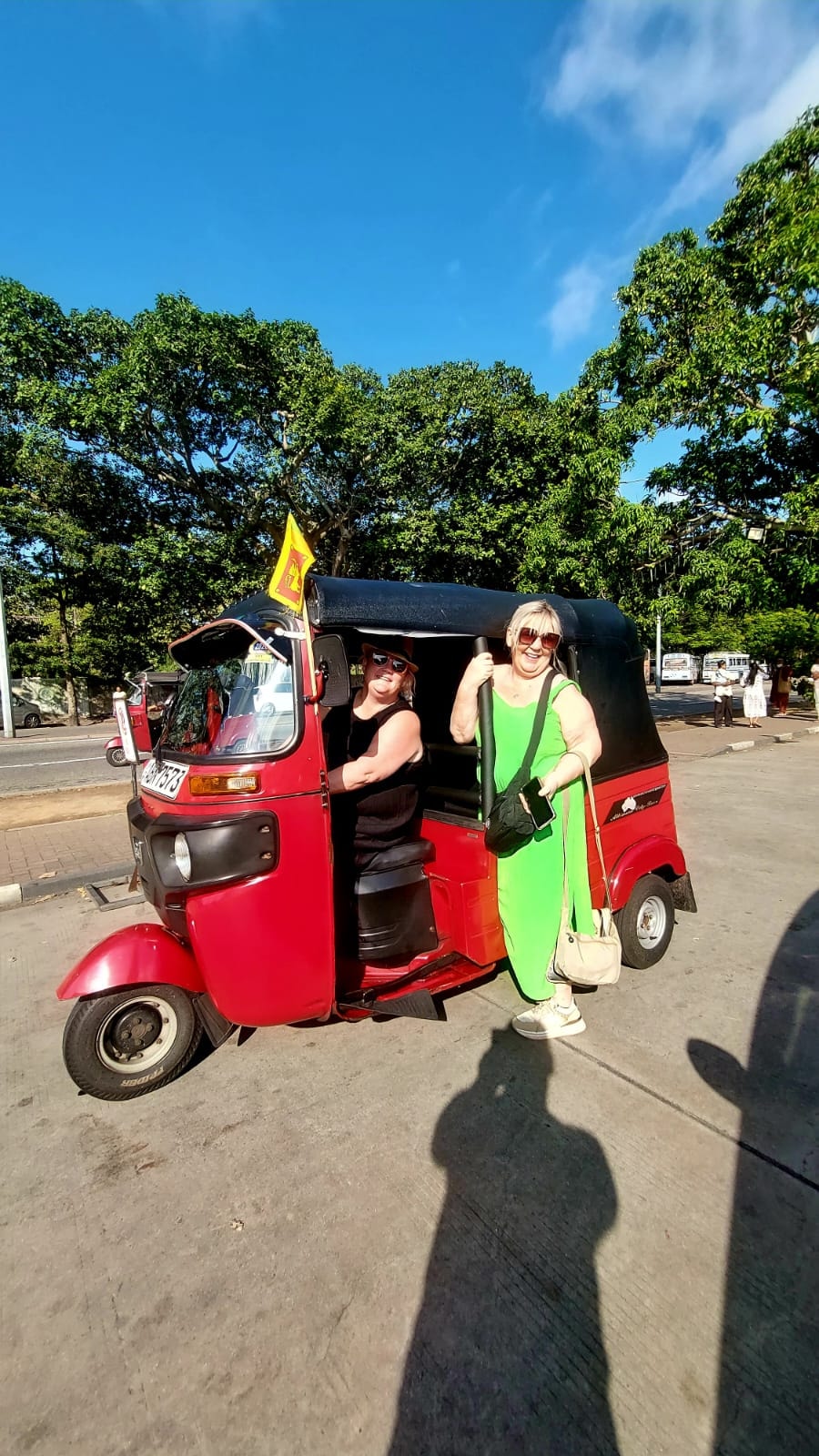 A guest beside a red tuk tuk on a sunlit tree-lined street